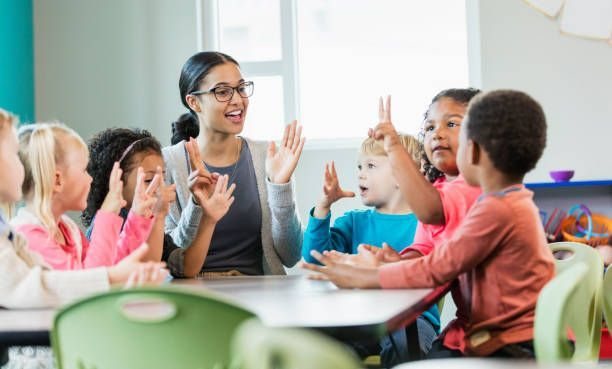 An adult and children raise their hands and make finger gestures around a table indoors.