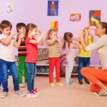Children stand in a group and copy hand movements demonstrated by a kneeling adult indoors.