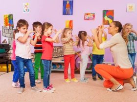 Children stand in a group and copy hand movements demonstrated by a kneeling adult indoors.