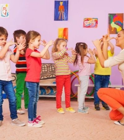 Children stand in a group and copy hand movements demonstrated by a kneeling adult indoors.