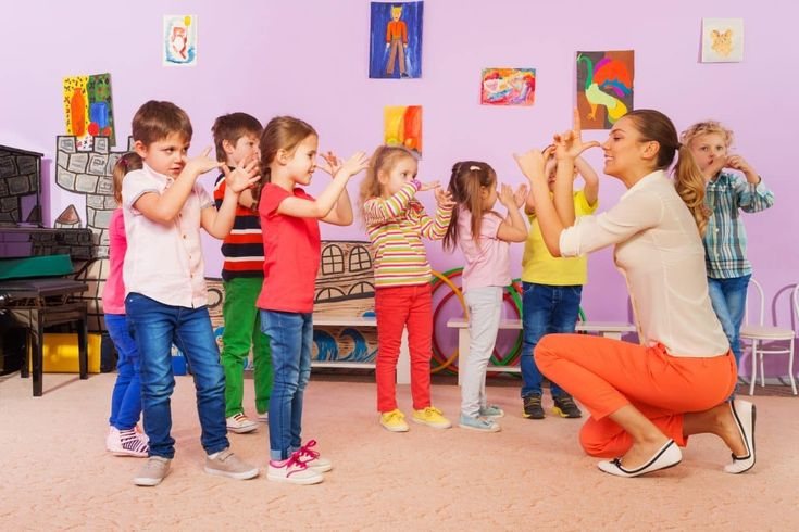 Children stand in a group and copy hand movements demonstrated by a kneeling adult indoors.